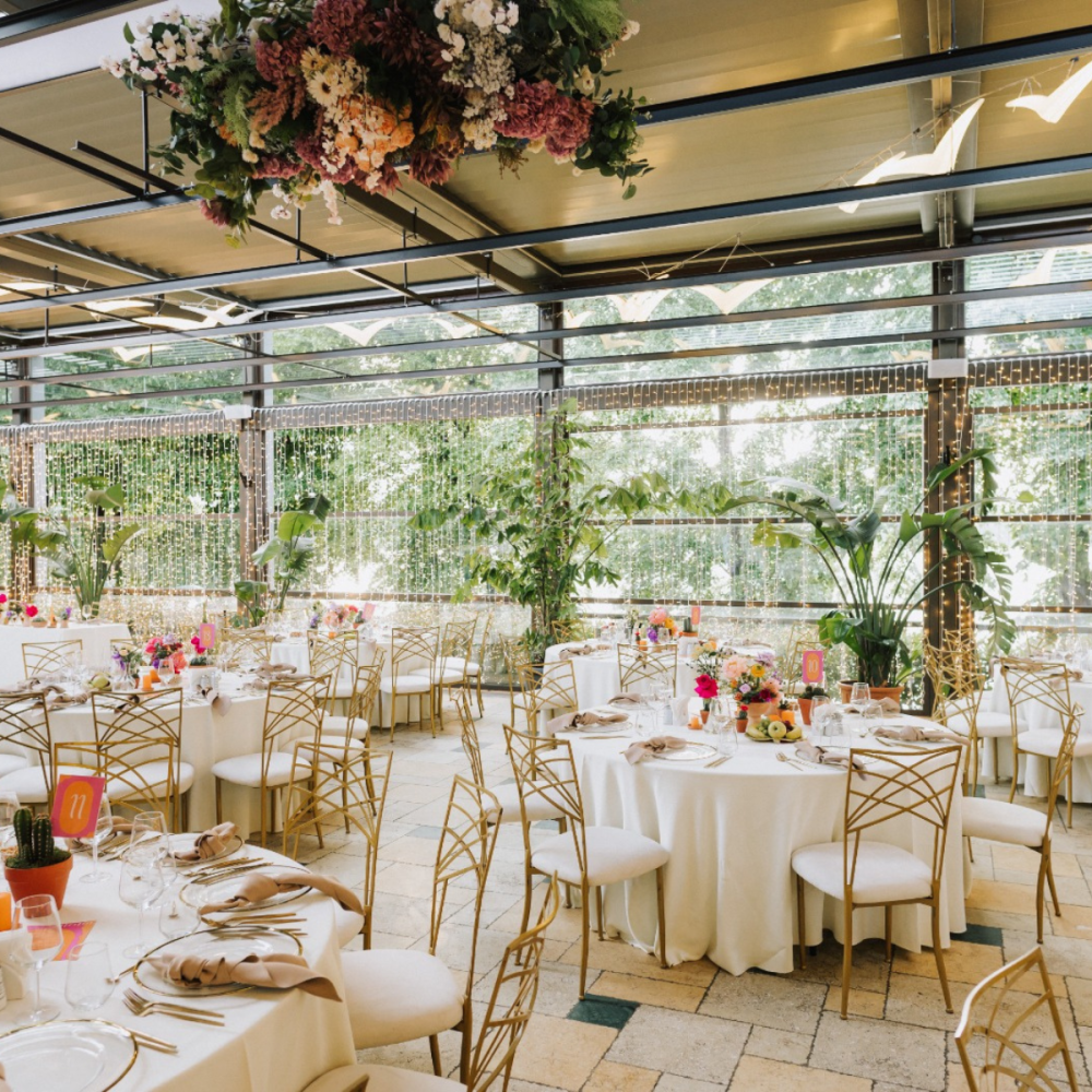 Elegant event reception in a glass-walled conservatory with round tables, white linens, gold chairs, and floral centerpieces.