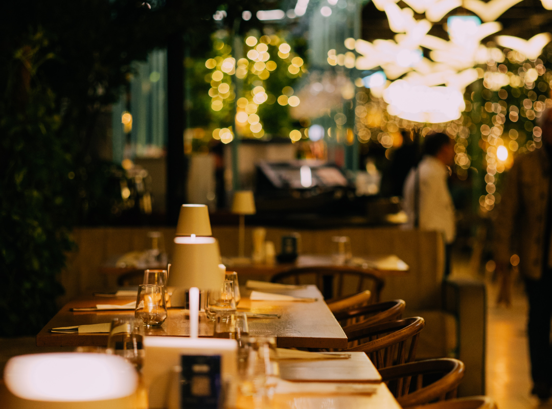 orangerie 2 - Restaurant Pescăruș Warmly lit restaurant interior with a long wooden table set with glasses, napkins, and small lamps; soft bokeh lights in the background.