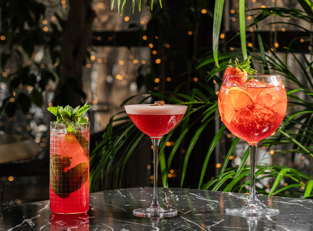 cocktails - Restaurant Pescăruș Three cocktails on a dark marble bar: a tall red drink with mint, a red coupe, and a large pink-red drink with a strawberry garnish.