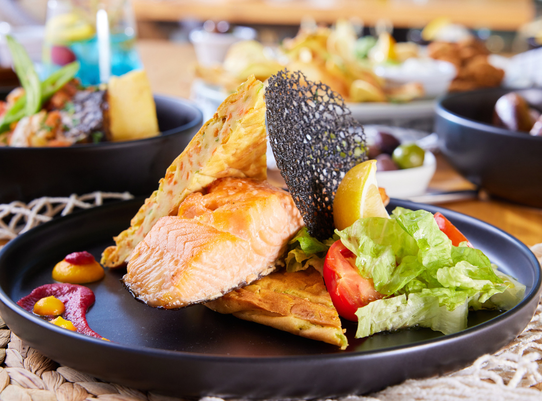 somon - Restaurant Pescăruș Pan-seared salmon on a black plate with a lace cracker, greens, tomato, lemon, and herb flatbread on the side.