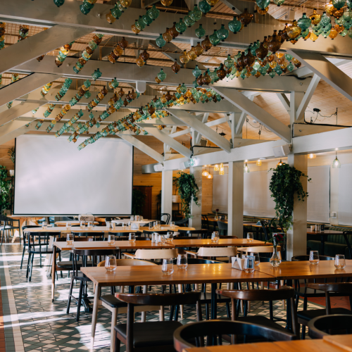 Event space in a restaurant with long wooden tables set for guests, white beams, and hanging teal‑amber glass pendant lights over head