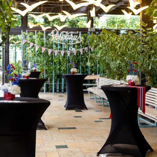 Decorated indoor event space with black cocktail tables, red-white-blue flower centerpieces, Union Jack bunting, and yellow bird-shaped lights.